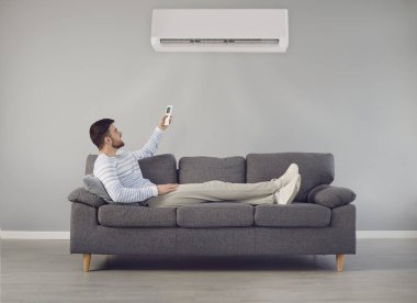 Genç adam odadaki kanepede otururken klimayı açar ve havayı soğutur.