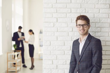 Beyaz bir ofiste, beyaz bir duvarda günlük kıyafetlerle gülümseyen pozitif iş adamı..