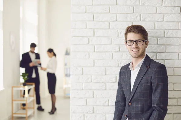 Leende positiv affärsman i avslappnad klädsel stående i ett vitt kontor på en vit vägg. — Stockfoto