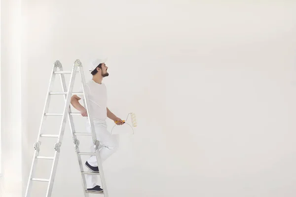 Um pintor masculino de uniforme branco com um rolo trabalha em sua mão em uma sala branca — Fotografia de Stock