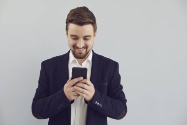 Ceketli olumlu adam gri arka planda gülümseyen bir cep telefonuyla metin yazıyor..