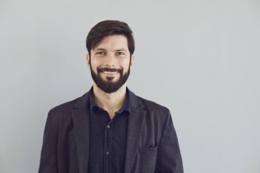 Ceketli olumlu sakallı adam gri arka plandaki kameraya bakıp gülümsüyor..