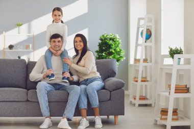 Mutlu gülümseyen aile. Kızı babasının omuzlarında, annesi de kanepede oturuyor.