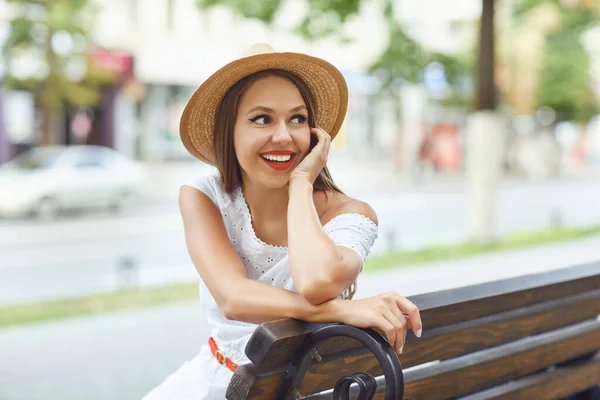 A kalapos lány mosolyog, egy padon ül a város utcáján.. — Stock Fotó