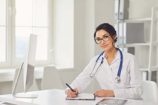 Confident woman doctor pediatrician writes in a clipboard sitting at a table in a white office of the hospital — Stok fotoğraf