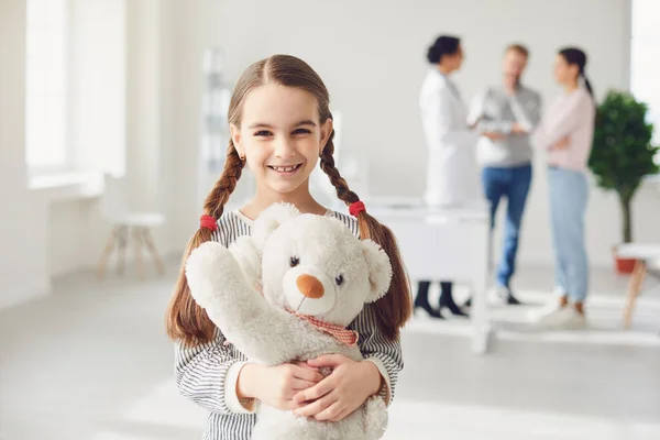 一个拿着玩具的女孩站在医生和站在诊所办公室的父母的背景下. — 图库照片