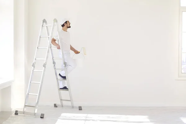 Un pintor masculino con un uniforme blanco con un rodillo trabaja en su mano en una habitación blanca — Foto de Stock