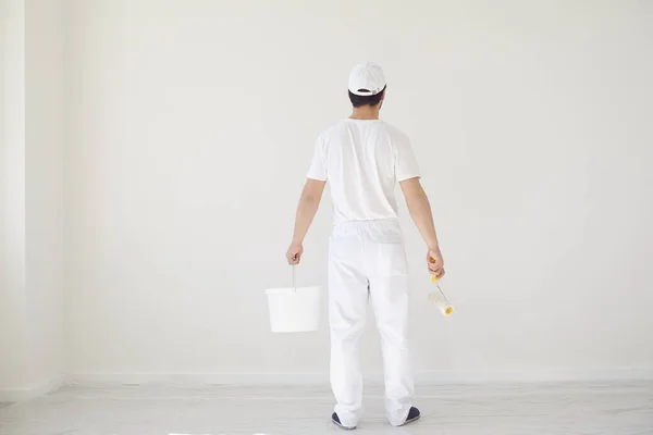Um pintor masculino de uniforme branco com um rolo trabalha em sua mão em uma sala branca — Fotografia de Stock
