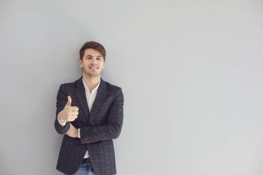 Gülümseyen adam gri bir arka planda sıradan bir ceketle başparmağını kaldırdı..