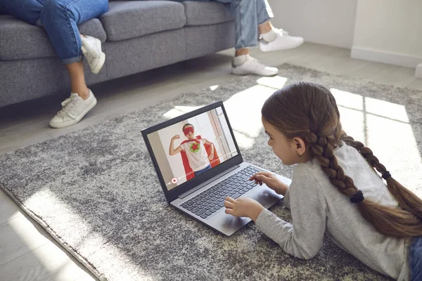 Menina criança assistindo online vídeo game entretenimento deitado confortavelmente no chão em uma sala com a família em casa . — Fotografia de Stock