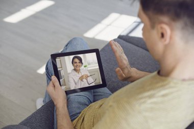 Doktor online konsepti. Bir adam evdeki kanepede otururken tablet kullanarak doktorla bağlantı kuruyor..