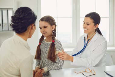 Kadın bir pediatrist, annesi klinikte olan bir kıza setoskop muayenesi yapıyor..