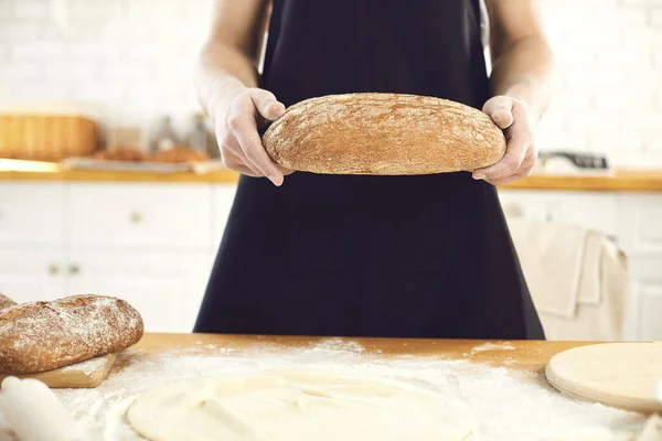 Человек-пекарь с бородой держит в руках свежий хлеб, стоящий на кухне. . — стоковое фото