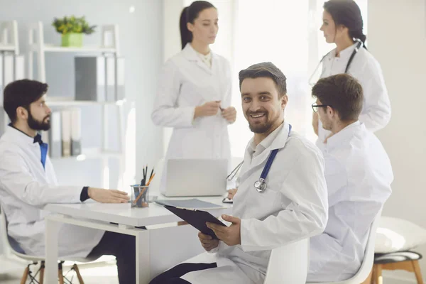 Medico maschio seduto a un tavolo con collegi in camici bianchi in un ospedale medico . — Foto Stock
