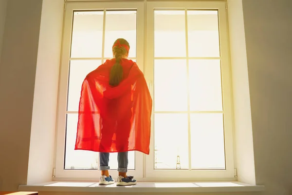Дитяча дівчина в костюмі супергероя, що стоїть на фоні вікна з сонячним світлом, дивлячись мрії на вікно — стокове фото