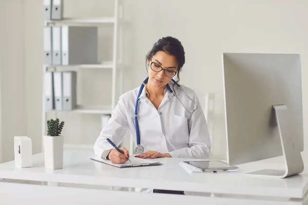 Confident woman doctor pediatrician writes in a clipboard sitting at a table in a white office of the hospital — 스톡 사진