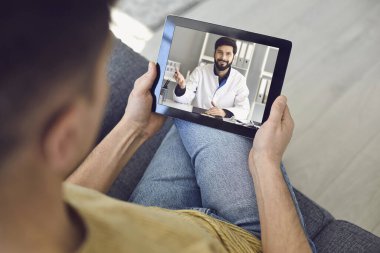 Hastalar için çevrimiçi tıbbi danışmanlık. Doktor Man, evdeki kanepede oturan erkek hastayla dizüstü bilgisayarla konuşuyor..