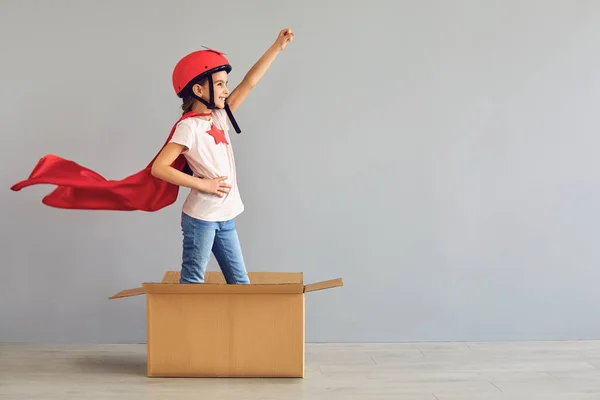 Дитина в костюмі супергероя підняв руку вгору, стоячи в картонній коробці сірий фон . — стокове фото