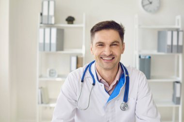 Steteskopla gülümseyen bir erkek doktor klinikte beyaz bir masada otururken..