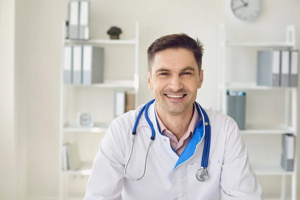 Mężczyzna lekarz z stetoskopem uśmiechnięty siedząc przy stole w białym wnętrzu gabinetu kliniki. — Zdjęcie stockowe