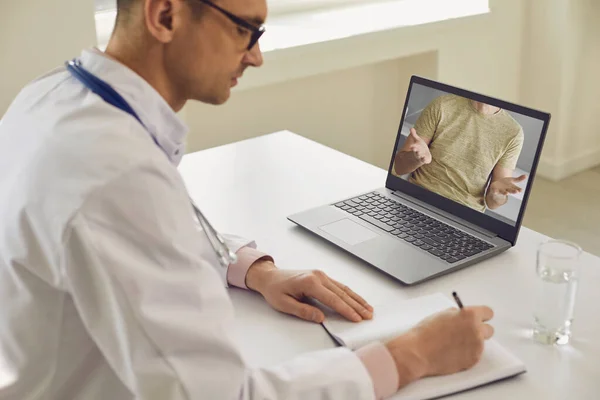 Médico en línea. El doctor habla con un paciente usando un portátil sentado detrás de una pajita en una clínica. Consulta médica en línea . — Foto de Stock