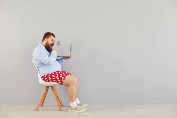 穿着红色短裤、手持笔记本电脑的有趣男人增加了灰色背景下的放大信息。在线自由职业者远程工作的概念. — 图库照片
