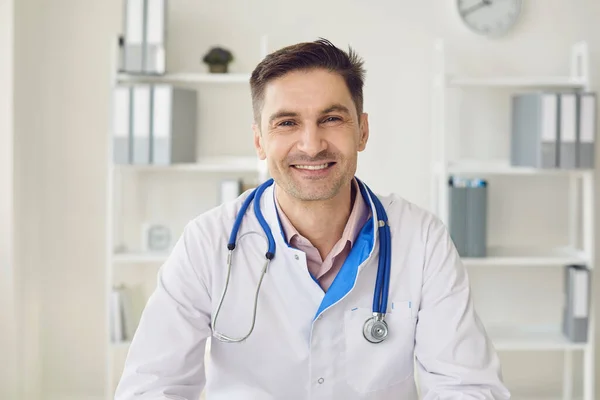 Médico varón con un estetoscopio sonriendo sentado en una mesa en un interior blanco de una clínica . — Foto de Stock