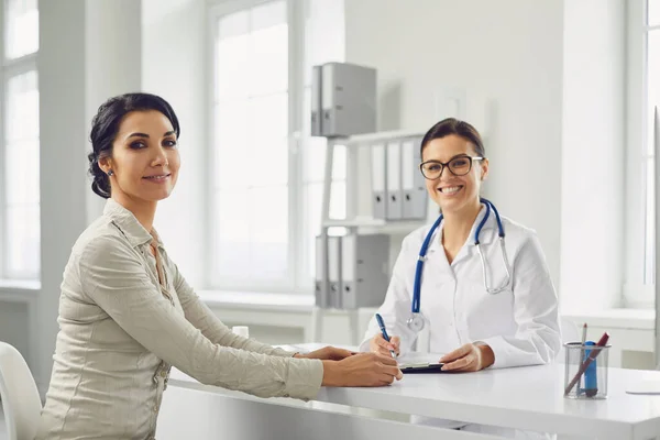 Paciente femenina sonriente en consulta con una doctora sentada en la mesa de la clínica del consultorio . —  Fotos de Stock