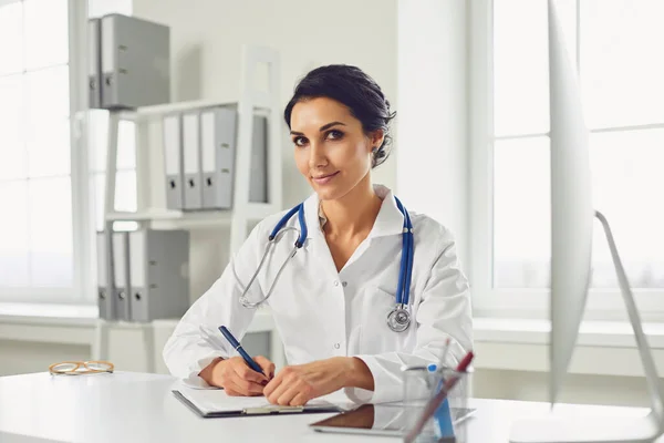 Confident woman doctor pediatrician writes in a clipboard sitting at a table in a white office of the hospital — Stok fotoğraf