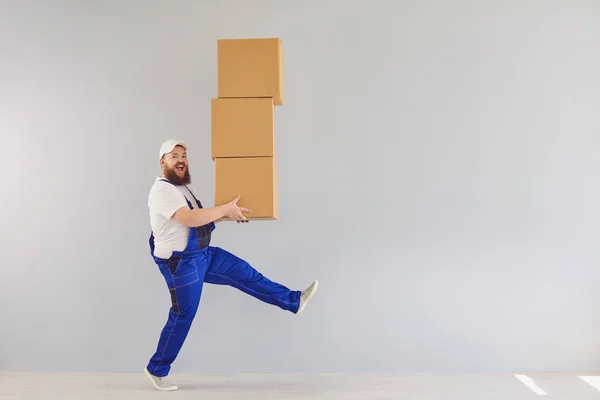 Funny fat bearded delivery courier loader man in a blue uniform with a cardboard box on a gray background.