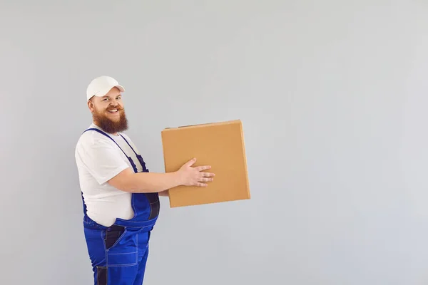 Funny fat bearded delivery courier loader man in a blue uniform with a cardboard box on a gray background.