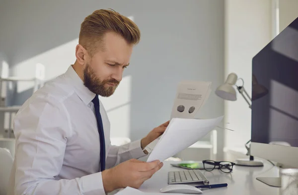 Un homme d'affaires sérieux et occupé lit des documents de travail assis à une table avec un ordinateur dans le bureau . — Photo