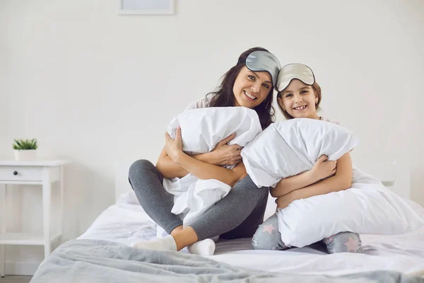 Glücklicher Muttertag. Mutter und Tochter in Schlafmasken lächeln, während sie mit Kopfkissen auf dem Bett im Zimmer in die Kamera schauen. — Stockfoto