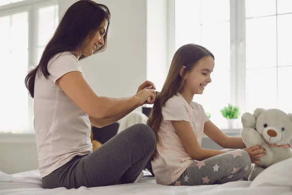 Ibu membuat gaya rambut untuk anak perempuan duduk di tempat tidur di kamar tidur. — Stok Foto