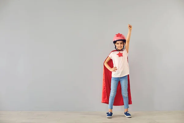 Little baby superhero in a helmet smiles raised his hands while standing against a gray background. — Stock Photo, Image