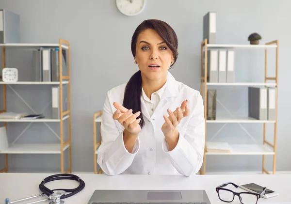 Internetowy lekarz konferencyjny patrzący w kamerę. Lekarz kobieta rozmawia z pacjentem wideo rozmowy na tle gabinetu kliniki. — Zdjęcie stockowe