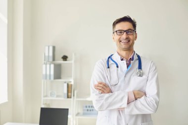 Beyaz önlüklü pozitif doktor kliniğin beyaz ofisinde kollarını kavuşturmuş duruyor..