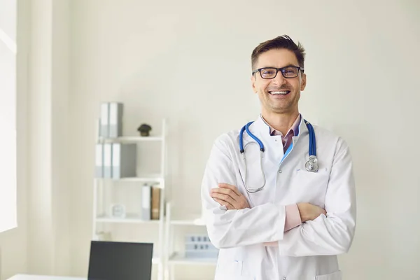 Pozytywny lekarz-terapeuta w białym płaszczu skrzyżował ramiona stojąc w białym gabinecie kliniki medycznej. — Zdjęcie stockowe