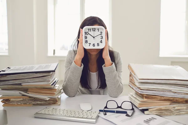 Mujer de negocios sosteniendo un despertador delante de su cara mientras está sentada en una mesa en la oficina — Foto de Stock