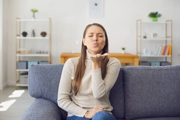 Young blonde girl gives an air kiss while sitting on a sofa in a living room at home. Online date at home. — Stock Photo, Image