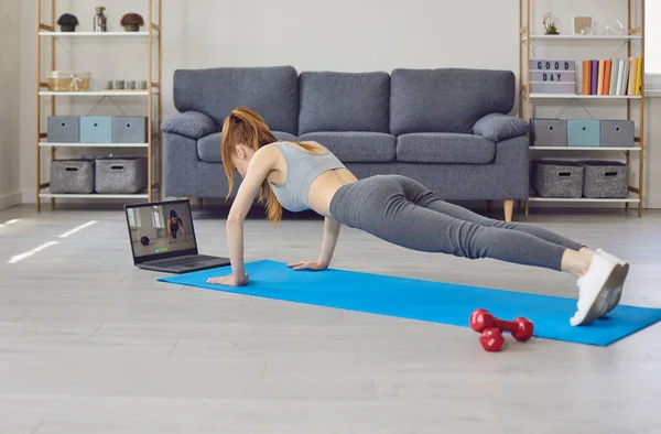 Trainen thuis online. Jonge vrouw doet push-ups uit te oefenen terwijl het kijken naar een laptop monitor thuis. — Stockfoto