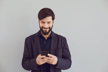 Ceketli olumlu adam gri arka planda gülümseyen bir cep telefonuyla metin yazıyor..