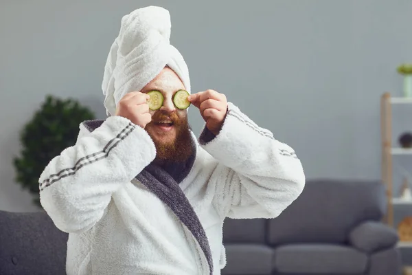 Странный толстый бородатый мужчина с косметической маской на лице в халате делает спа-процедуры на лице покоится в комнате дома — стоковое фото