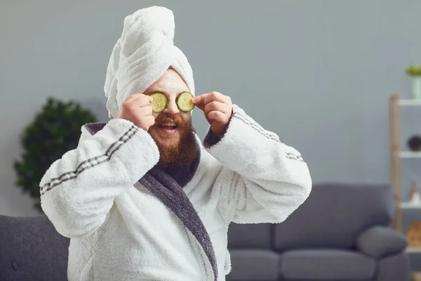 Divertente strano grasso barbuto uomo con una maschera cosmetica sul viso in accappatoio fa trattamenti termali sul suo viso riposa in una stanza a casa — Foto Stock