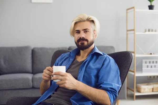 Online video chat. A young man with a mug of coffee looks at the camera in a living room.