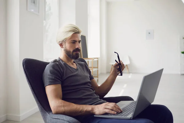 Pracuj online doma. Online školení. Mladý podnikatel v neformálním oblečení pracuje s notebookem, zatímco sedí v křesle v obytném pokoji. — Stock fotografie