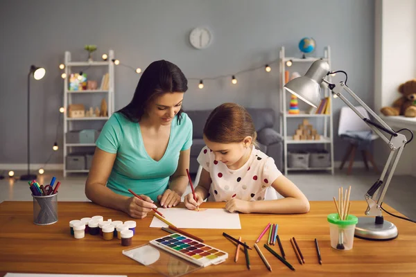 Щаслива сім'я. Маленька дівчинка і мати малюють на столі в кімнаті. Мама вчить доньку малювати, сидячи за столом удома.. — стокове фото