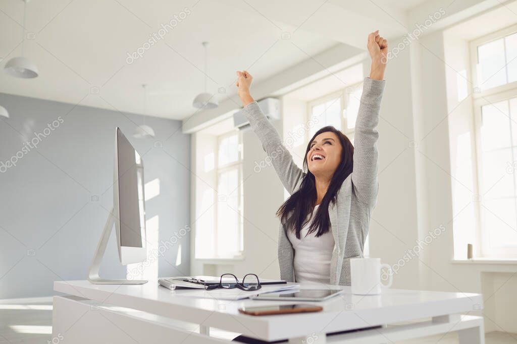 Happy successful smiling business woman raised her hands up while sitting at a table in the office.