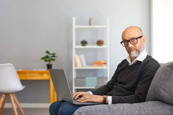 Arbeiten Sie zu Hause online. Schwerwiegender Mann mittleren Alters grauen Bart mit Laptop online arbeiten sitzt in einem Sessel in einem Raum. — Stockfoto