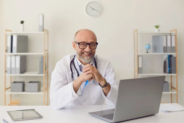 Medico maturo.Un medico fiducioso di mezza età sorride mentre siede a un tavolo con un computer portatile sul posto di lavoro nell'ufficio della clinica . — Foto Stock